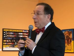 A man in a black suit jacket, red bowtie and white shirt holding a microphone.