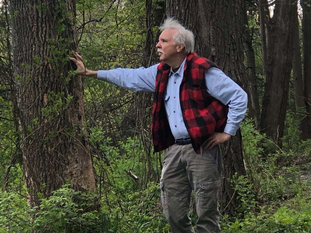 A man in a red plaid vest with a blue shirt and white hair in profile leaning against a tree.