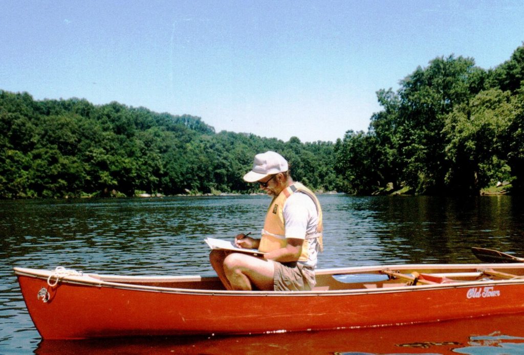 A man in a red canoe and wearing a faded yellow life jacket and a white cap writing in a spiral notebook.